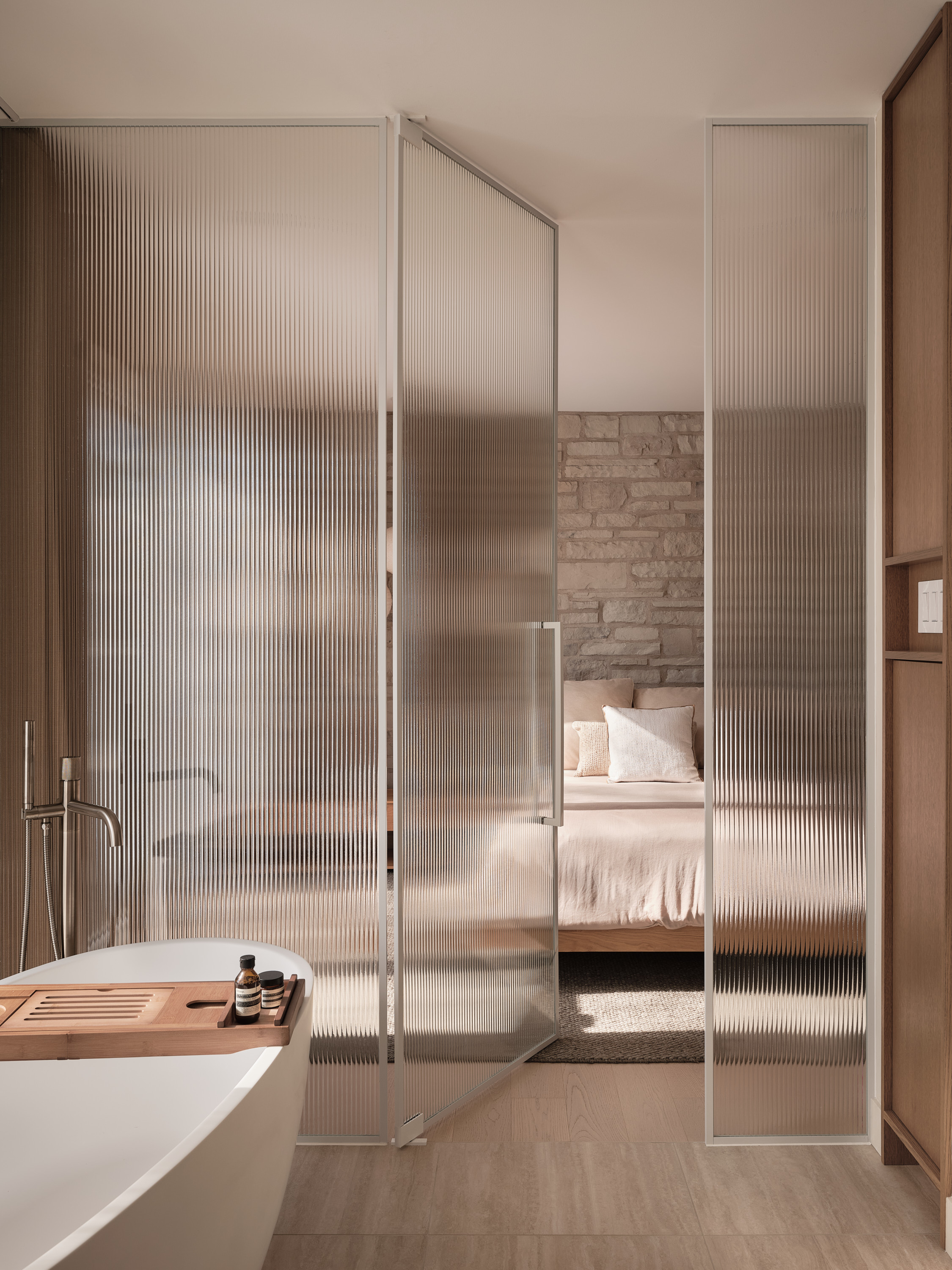 Freestanding bathtub with a wooden shelf, in front of a fluted glass partition opening onto the bedroom