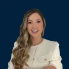 Smiling woman with long wavy hair wearing a white blazer against a dark blue background.