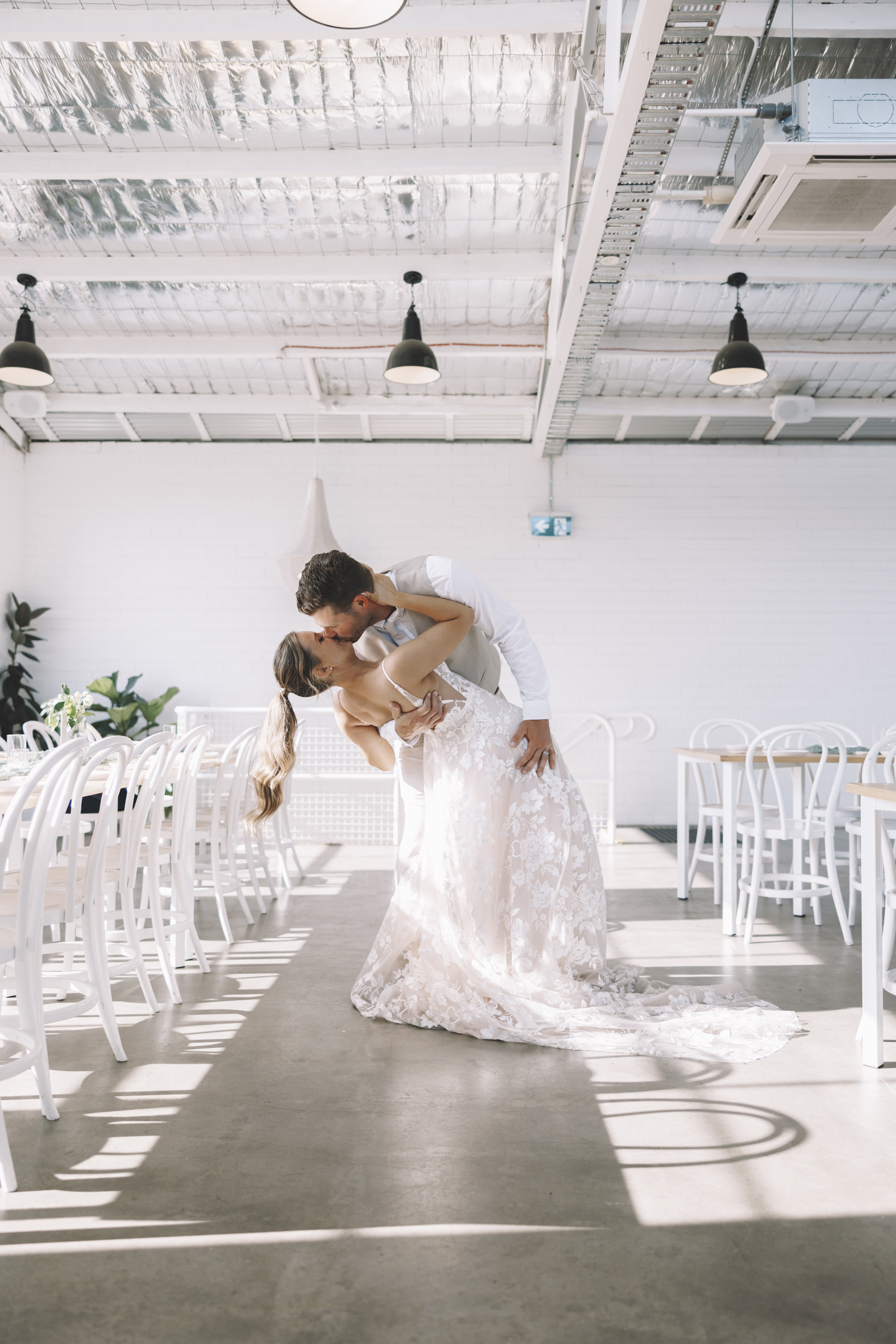 A groom dipping and kissing his bride in a sunlit white reception room