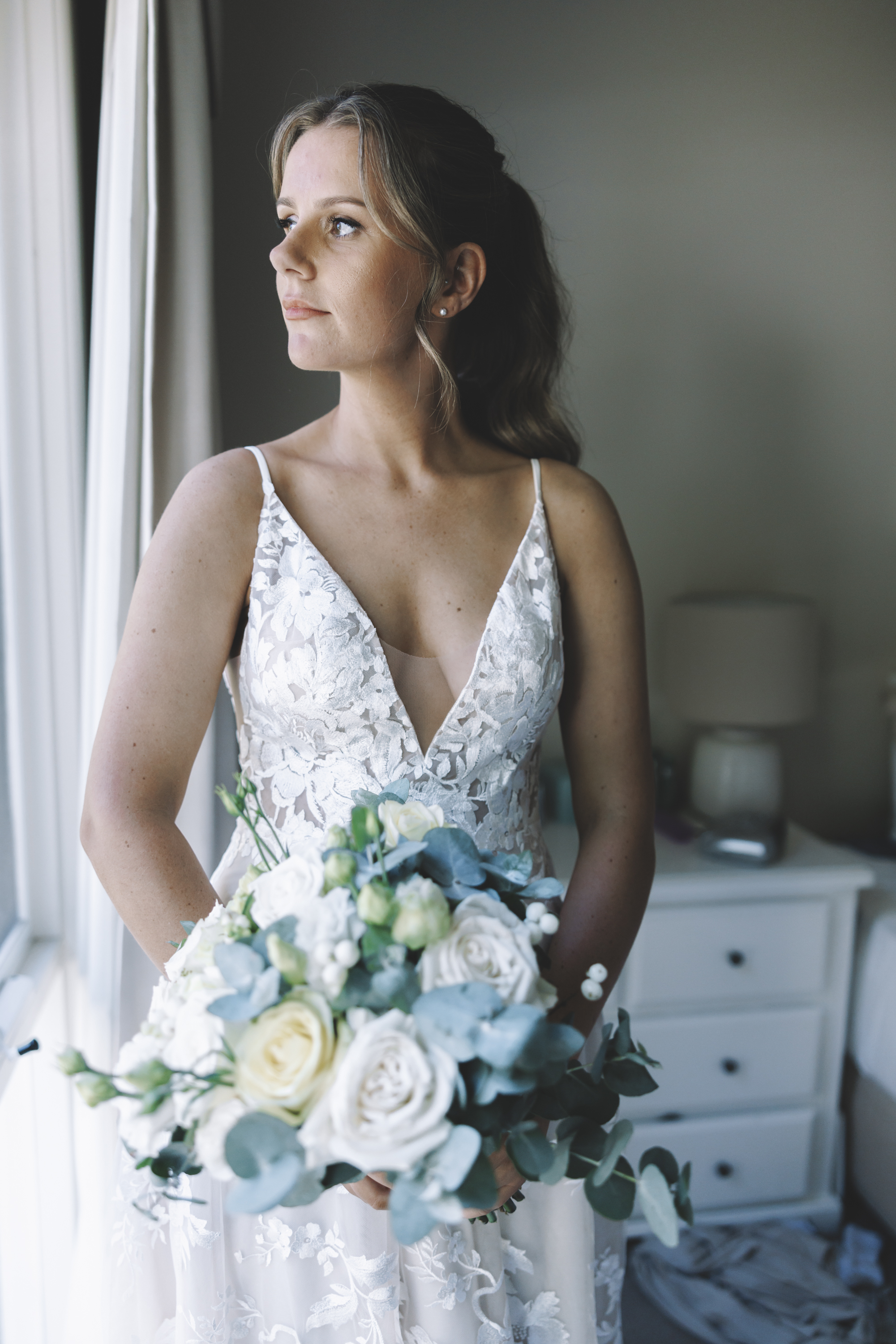A bride in a lace gown holding a eucalyptus and rose bouquet by a window