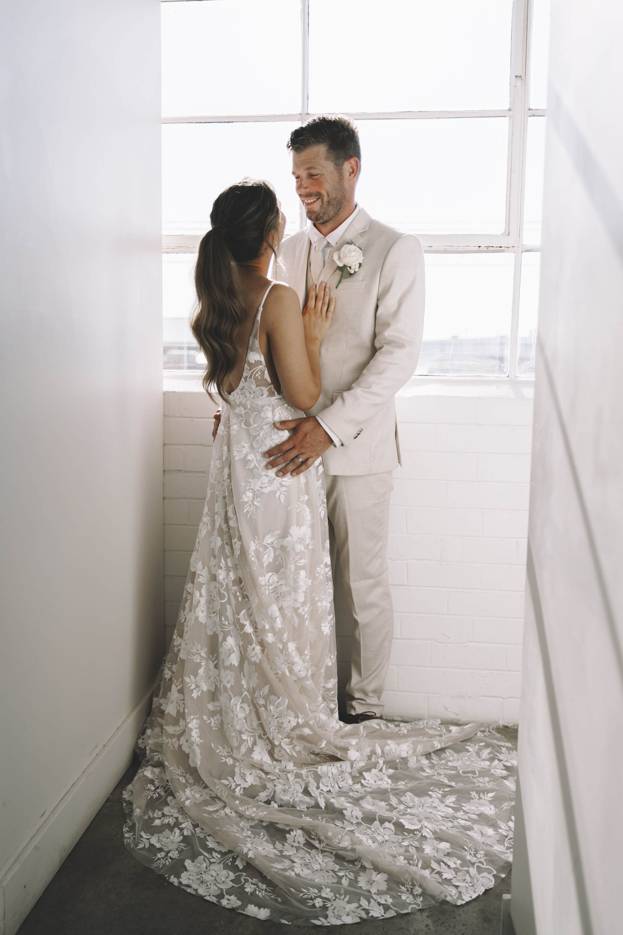 A smiling couple holding each other beside a tall, bright window