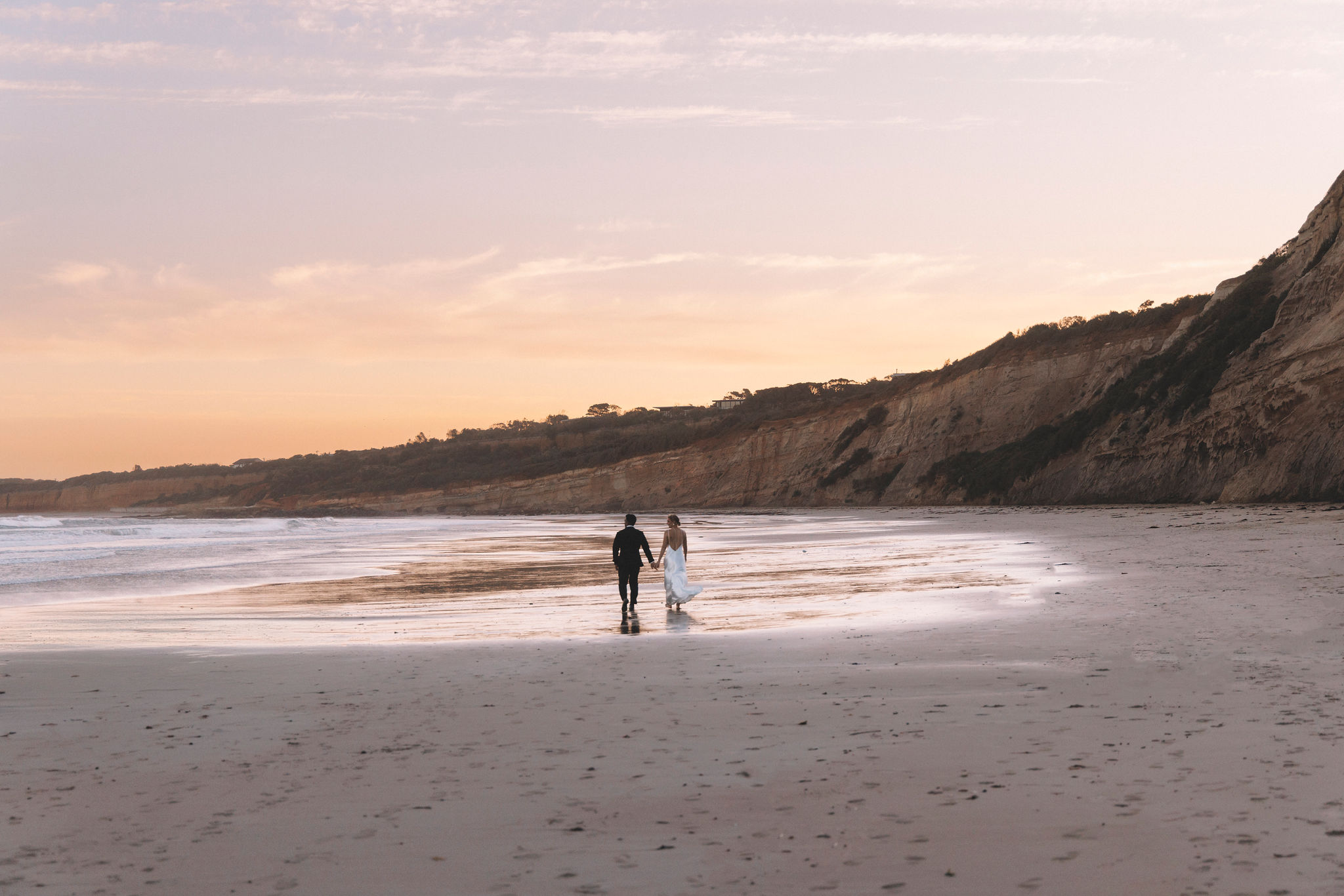 A couple walking hand in hand along the beach beneath sandstone cliffs at sunset