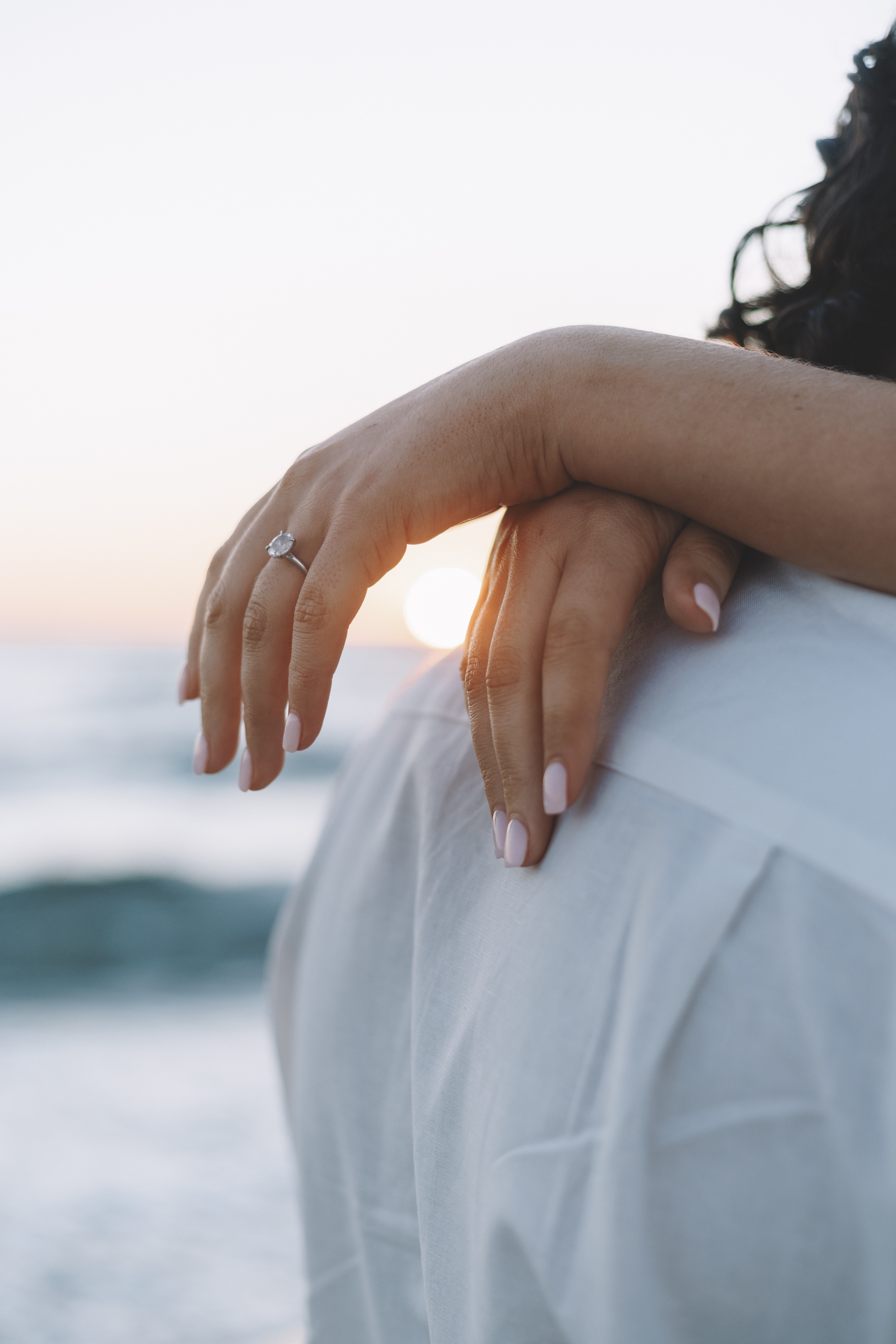 A hand wearing an engagement ring resting on a partner's shoulder by the sea