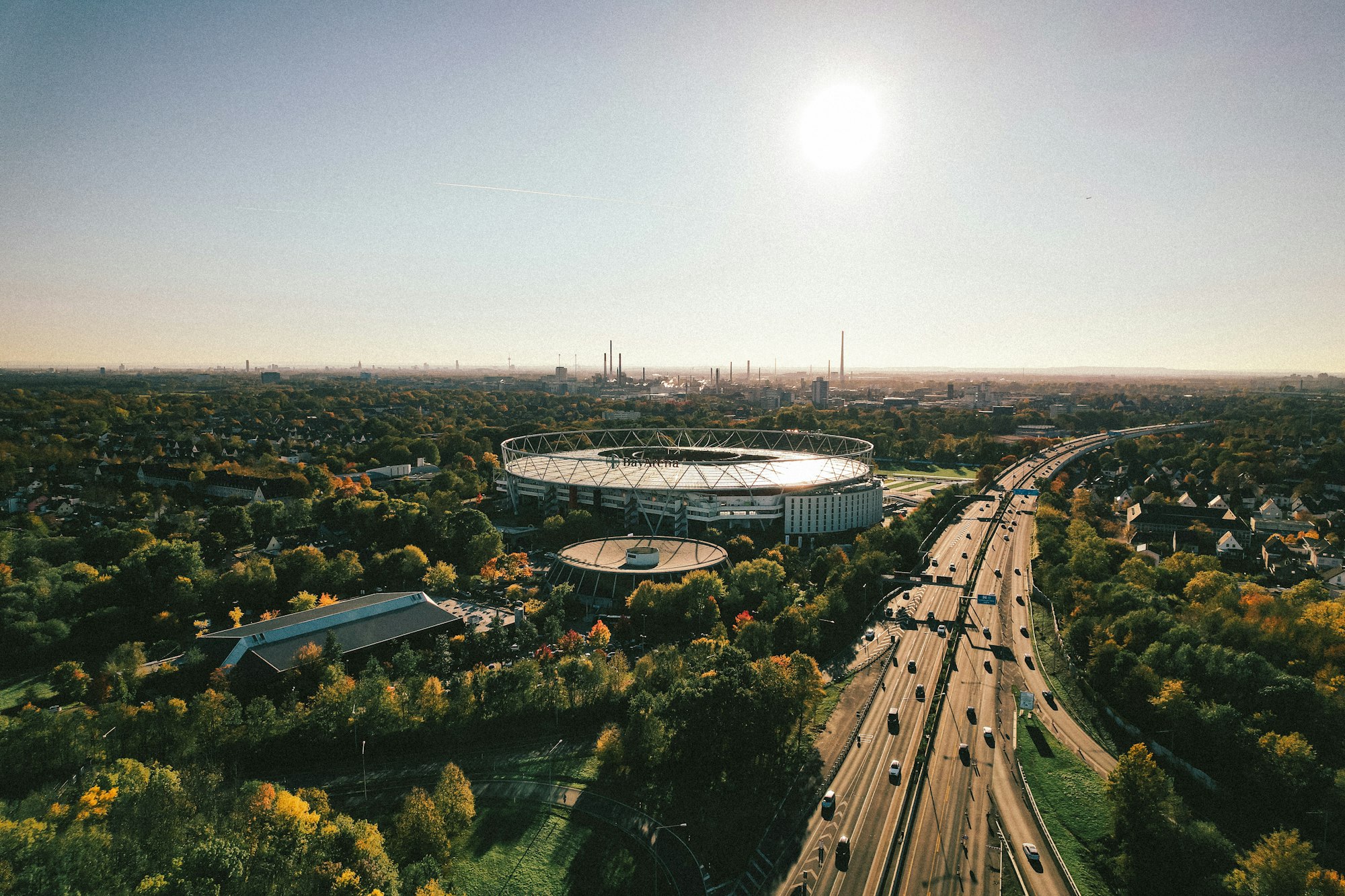 Vue aérienne de la BayArena et de son toit circulaire au milieu des arbres, à Leverkusen, avec une autoroute et le panorama de la ville à l'horizon