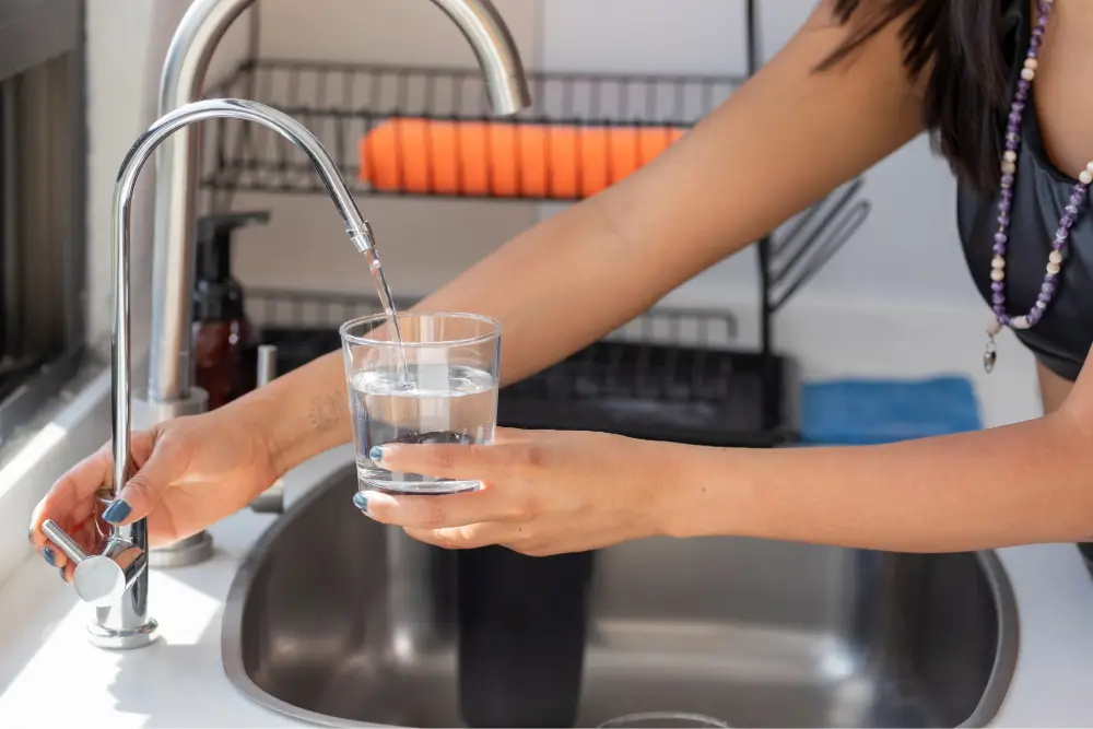 A person filling a glass with water from a modern kitchen sink faucet, with a dish rack and soap dispenser in the background.