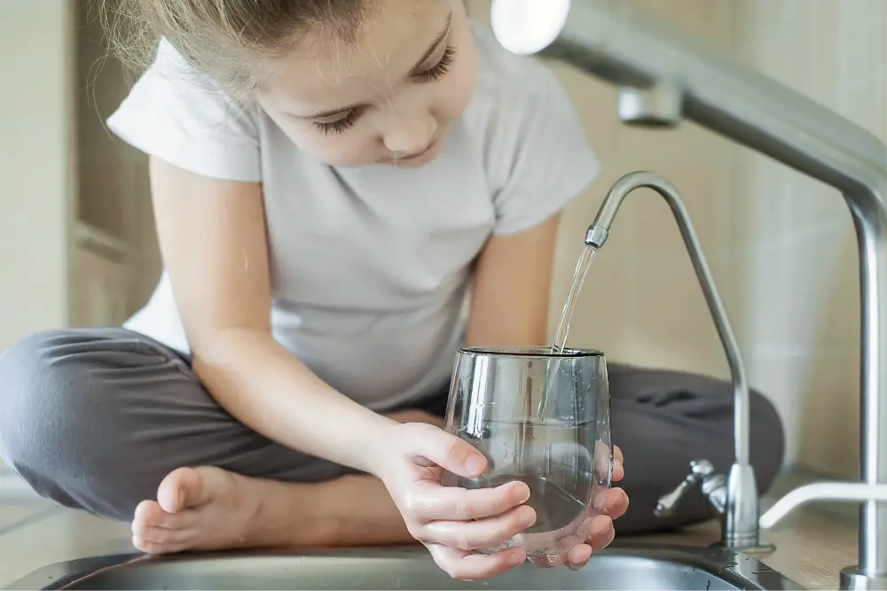 A young girl sitting cross-legged on a kitchen counter, holding a glass under a faucet to fill it with water.