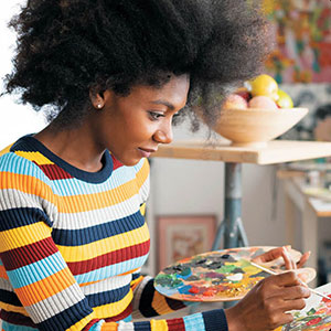 woman in striped shirt holding paint palette in left hand while painting with right hand