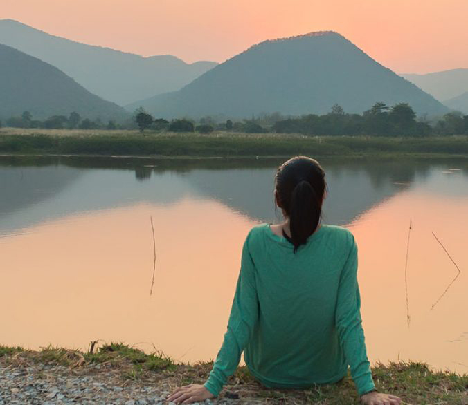 contemplative woman sitting by water at sunrise while gazing at mountains in the distance