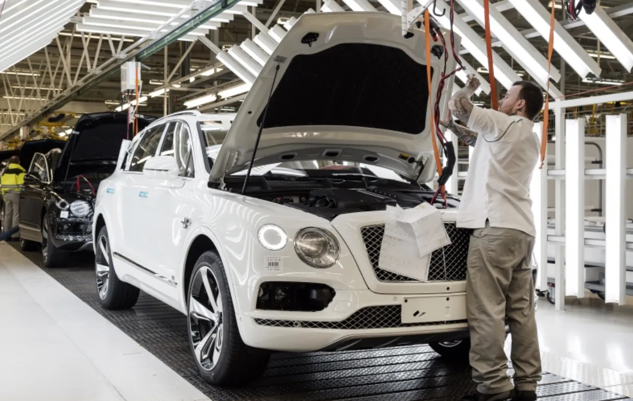 A factory worker assembling a white Bentley Bentayga SUV on a production line, with its hood open and cables connected. Other vehicles and bright overhead lights are visible in the background.