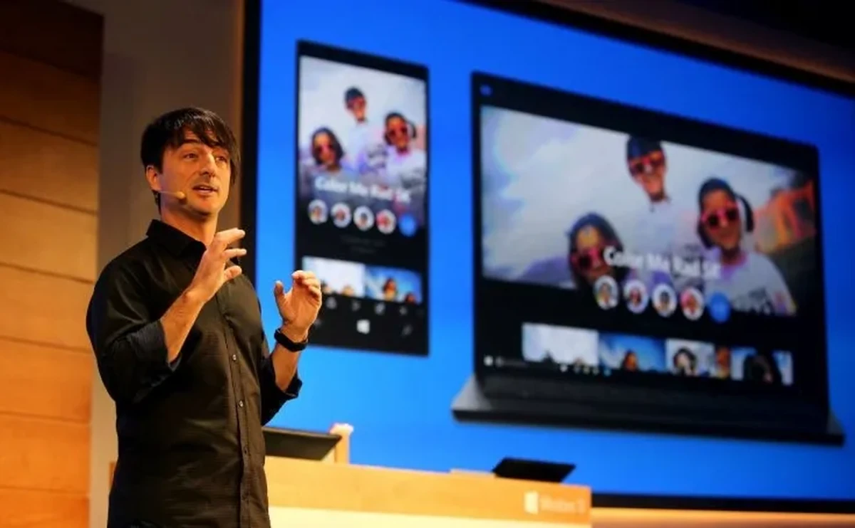 A person is presenting on stage in front of a display screen showing a smartphone and laptop. The screen features an app with a photo editing interface. The presenter is gesturing with hands while speaking.