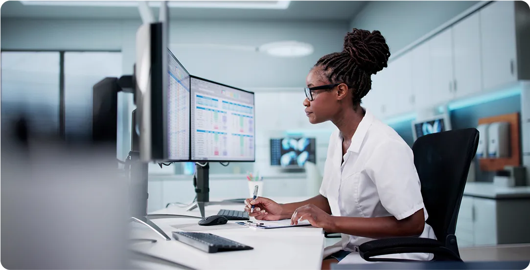 Healthcare professional reviewing data on dual computer monitors and taking notes in a clinical office.