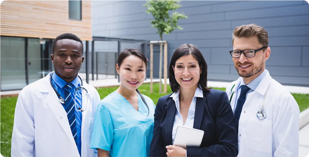 Four diverse healthcare professionals smiling outdoors, including two doctors in white coats, a nurse in blue scrubs, and a woman in a business suit holding a tablet.