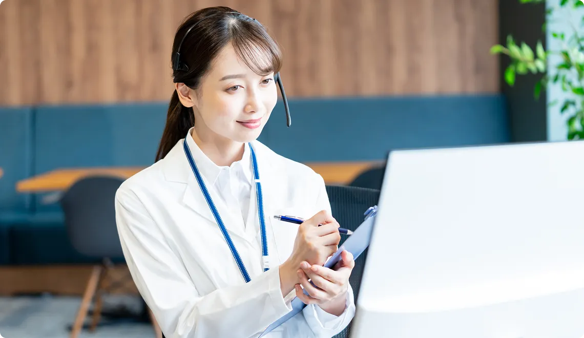Smiling female healthcare professional wearing a white coat and headset, holding a clipboard and pen, working at a computer.