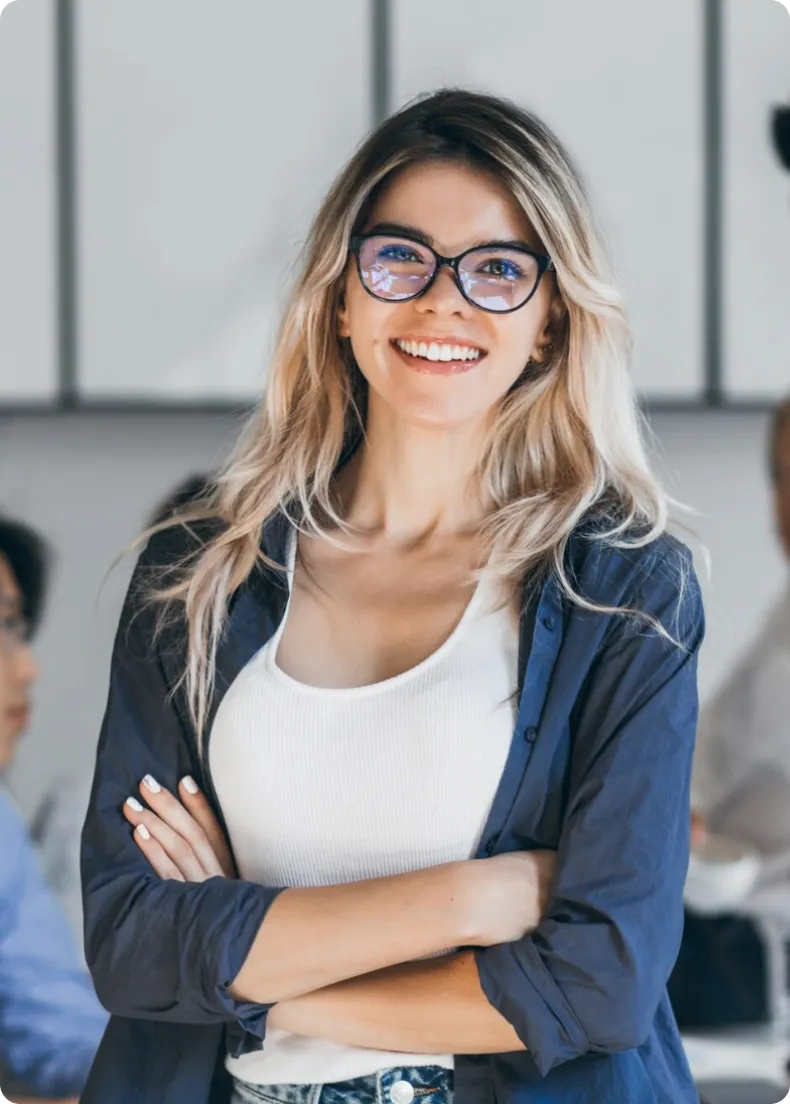 Smiling young woman with glasses, long blonde hair, wearing a white tank top and navy shirt, standing with arms crossed indoors.