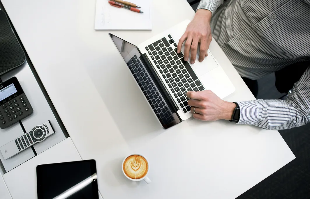 Overhead view of a person typing on a laptop at a white desk with a cup of coffee, tablet, calculator, and remote control nearby.