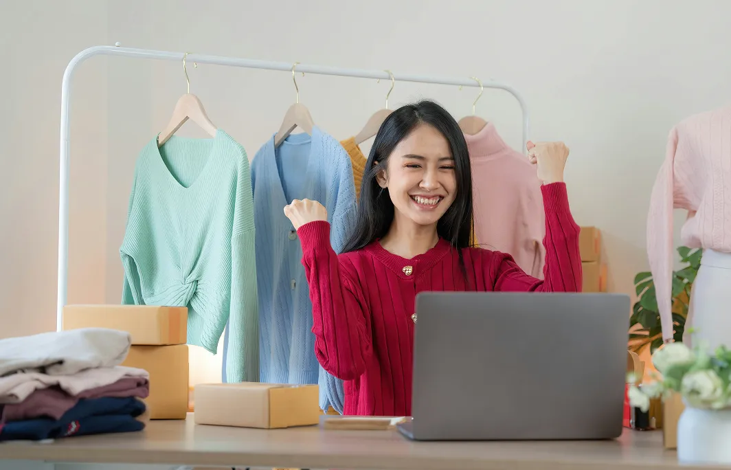 Smiling woman in a red sweater celebrating in front of a laptop with colorful sweaters hanging behind her and folded clothes on the table.