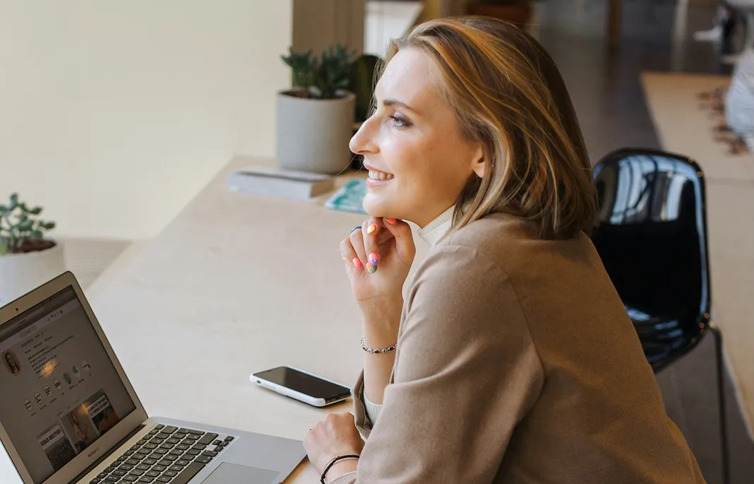 Smiling woman with colorful nails looking at a laptop screen while sitting at a desk with a smartphone and potted plants.