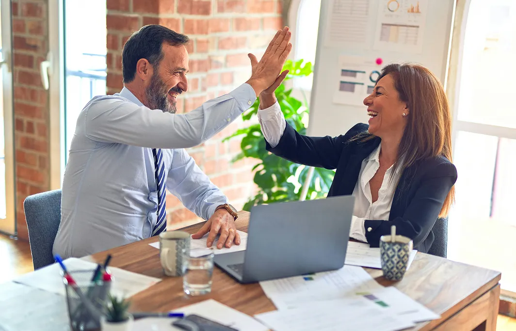 Two business colleagues smiling and giving each other a high five at a desk with a laptop and documents.