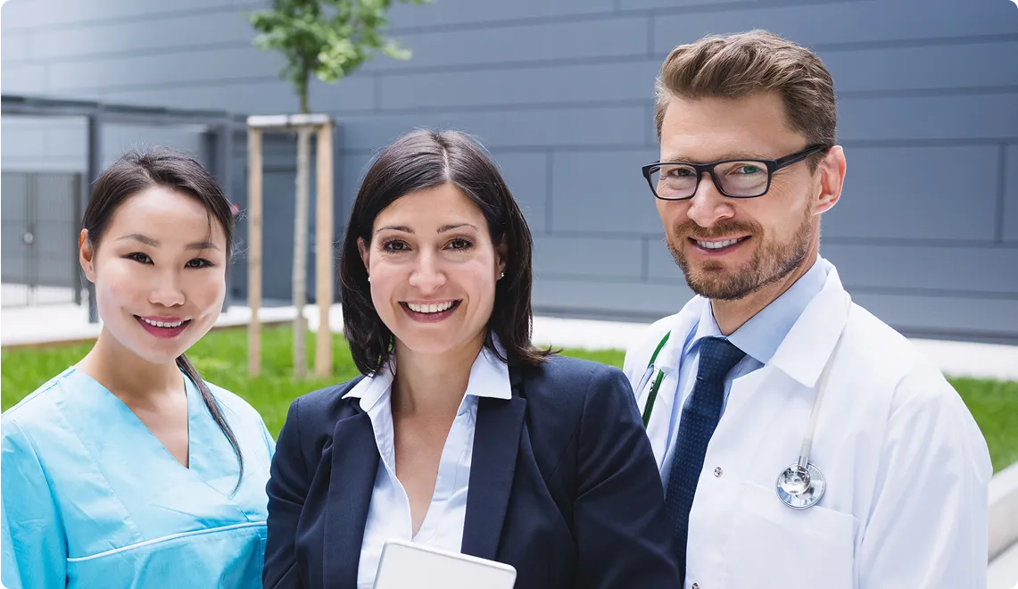 Three smiling healthcare professionals standing outdoors with a modern building wall in the background.
