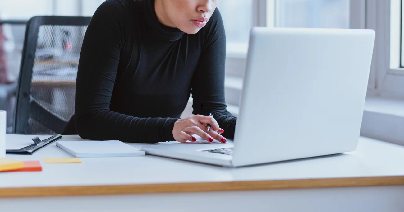 Woman in black long-sleeve top working on a silver laptop at a white desk with notebooks and sticky notes.
