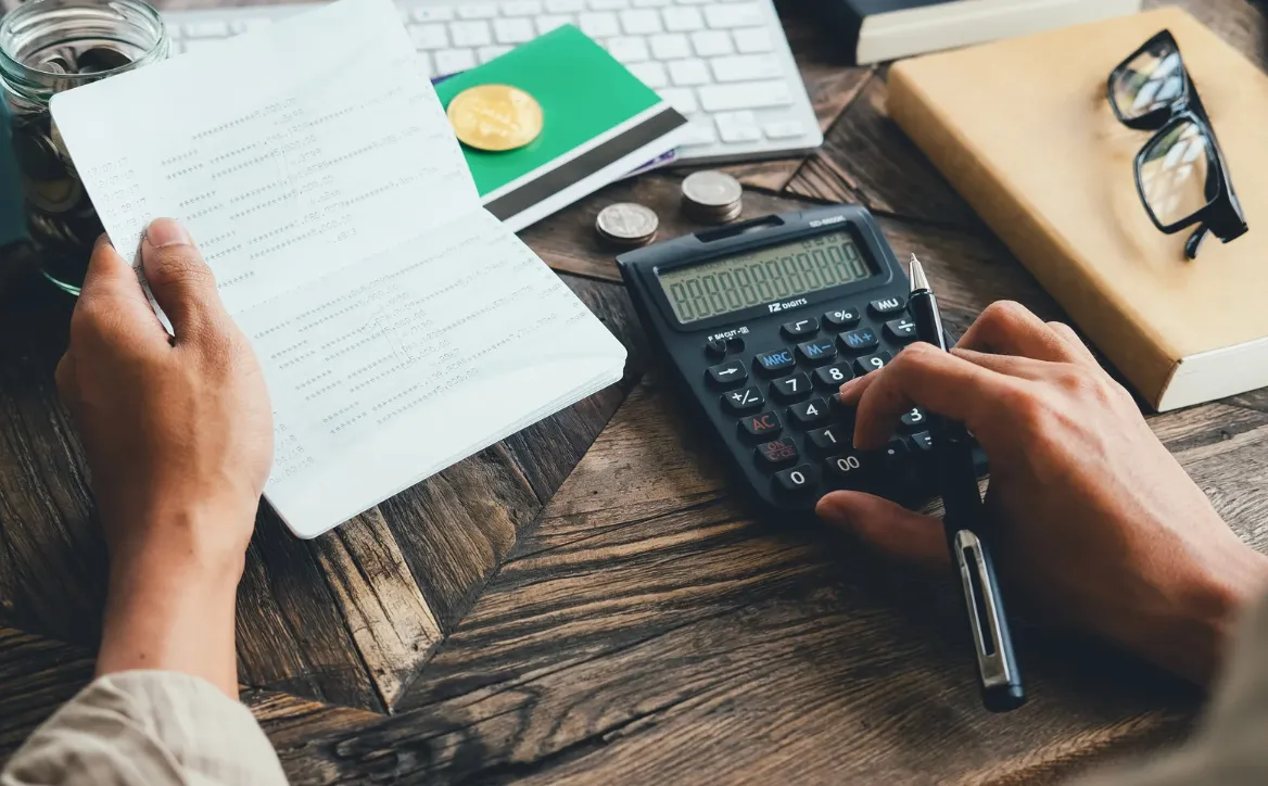 Person calculating expenses using a calculator and holding a printed receipt on a wooden desk with coins, glasses, and a book nearby.