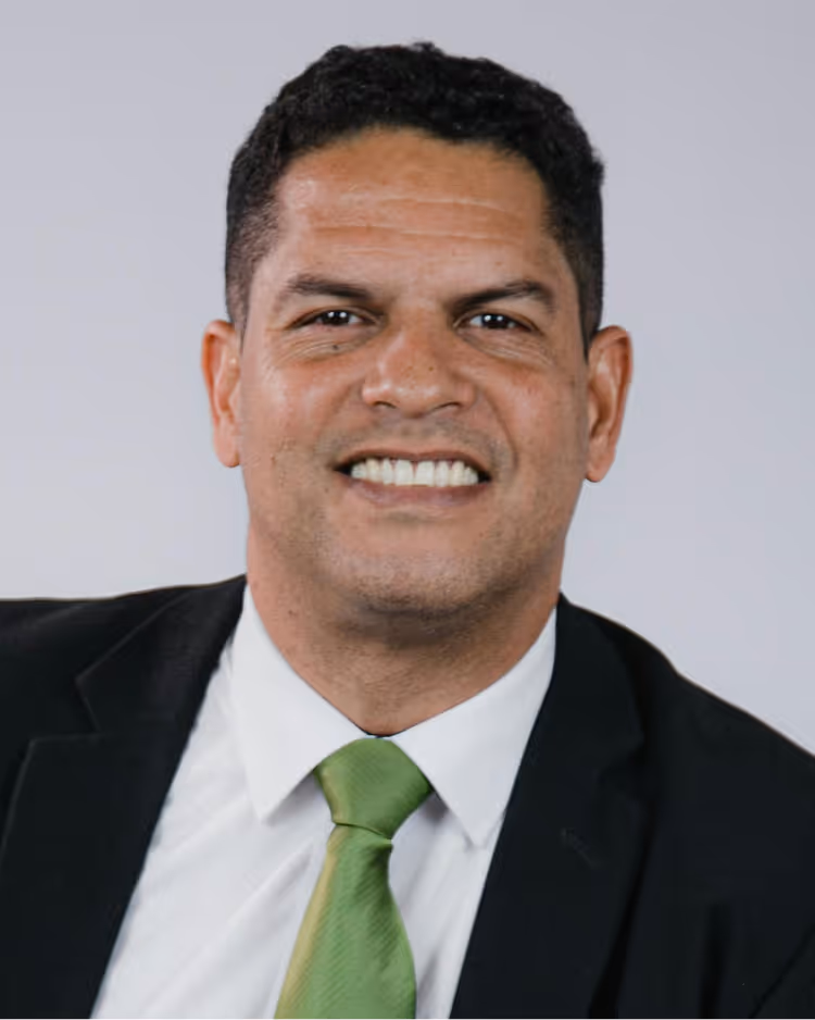 Smiling man with short black hair wearing a black suit, white shirt, and green tie against a plain light background.