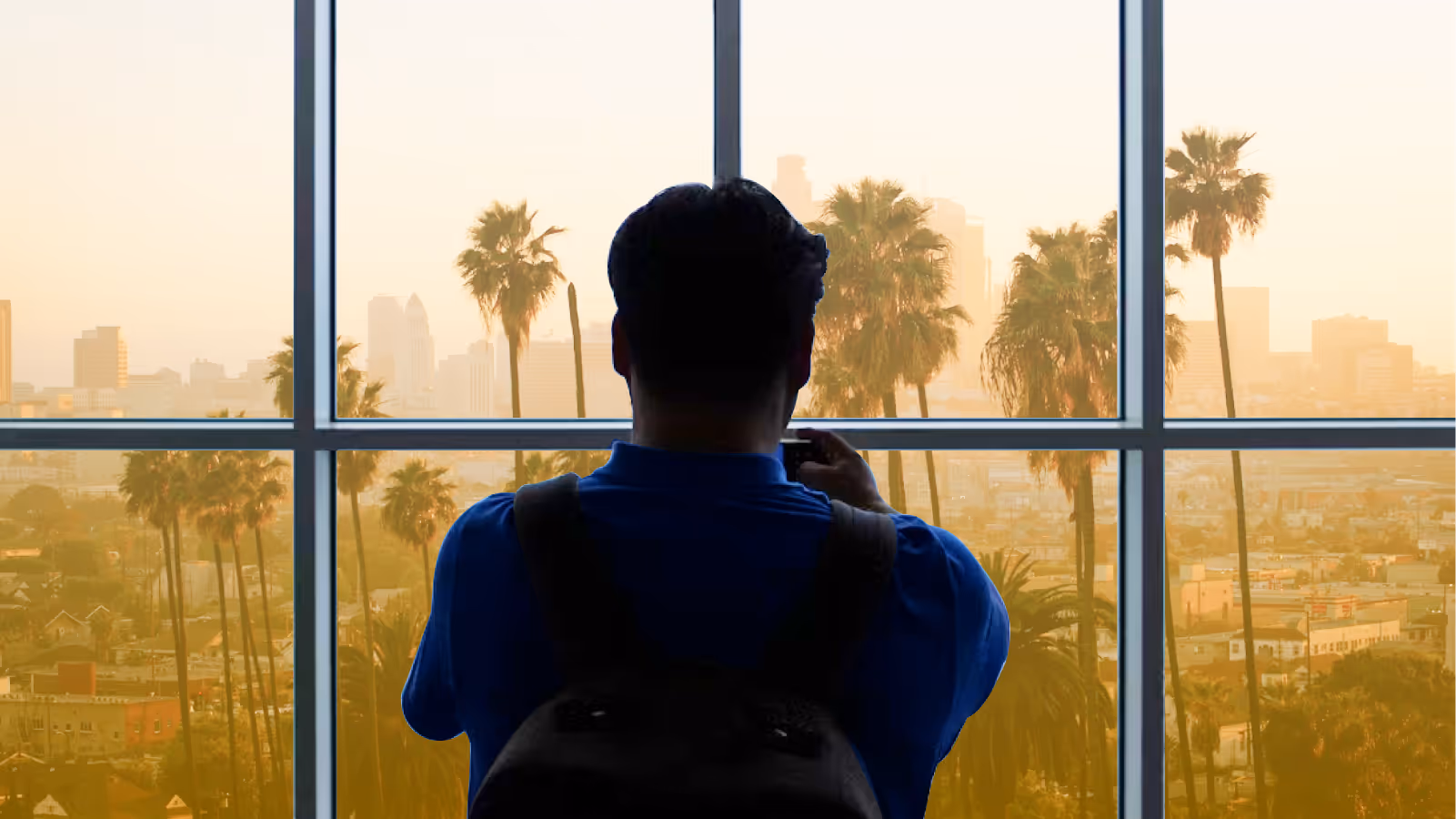 Person in blue shirt with backpack taking a photo through a large window overlooking a cityscape with palm trees at sunset.