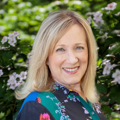 Blonde woman smiling in front of green foliage and pink flowers outdoors.