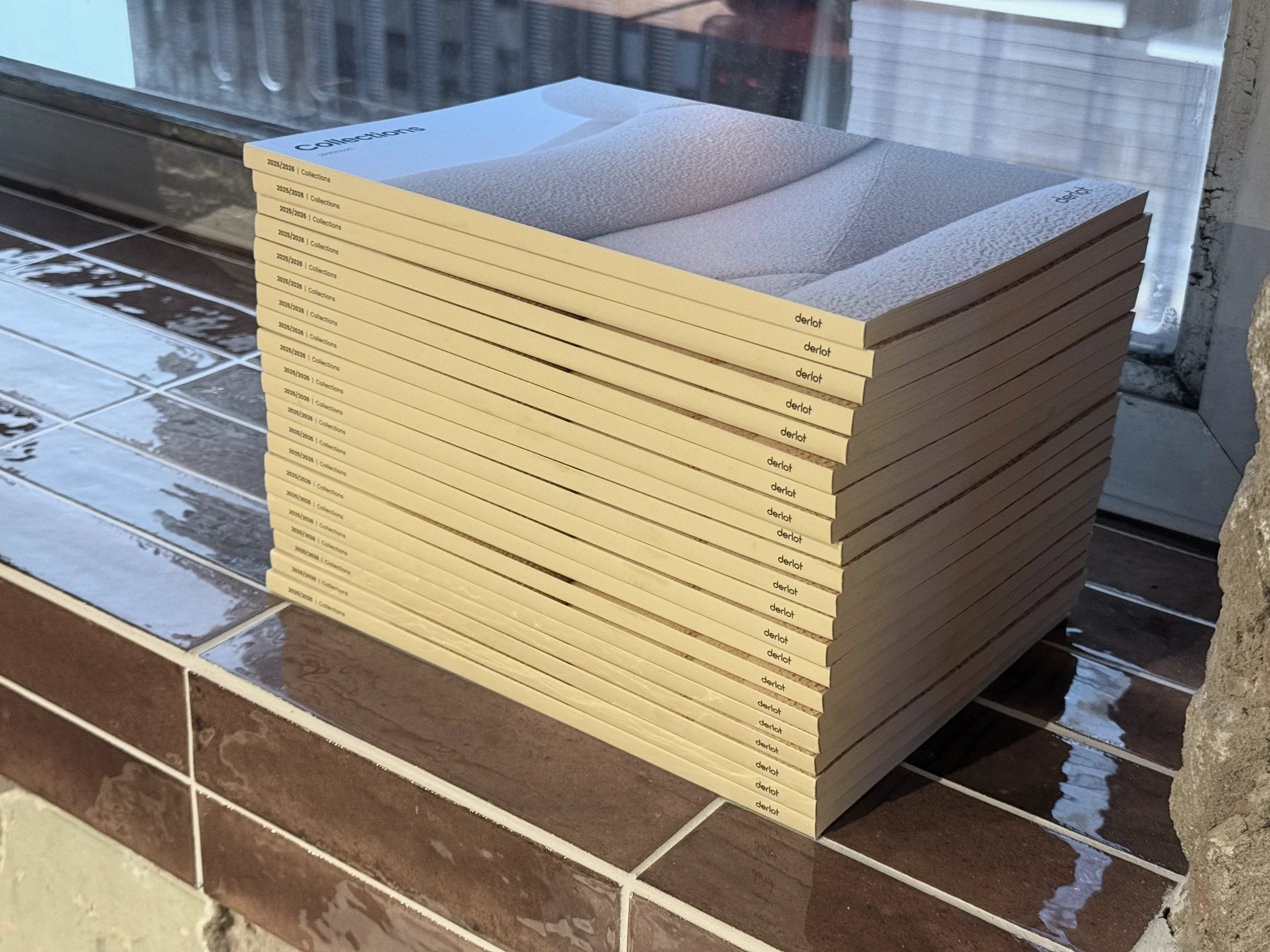 Stack of 20 catalogue books titled 'Collections 2025/2026' by Derlot on a brown tiled surface.