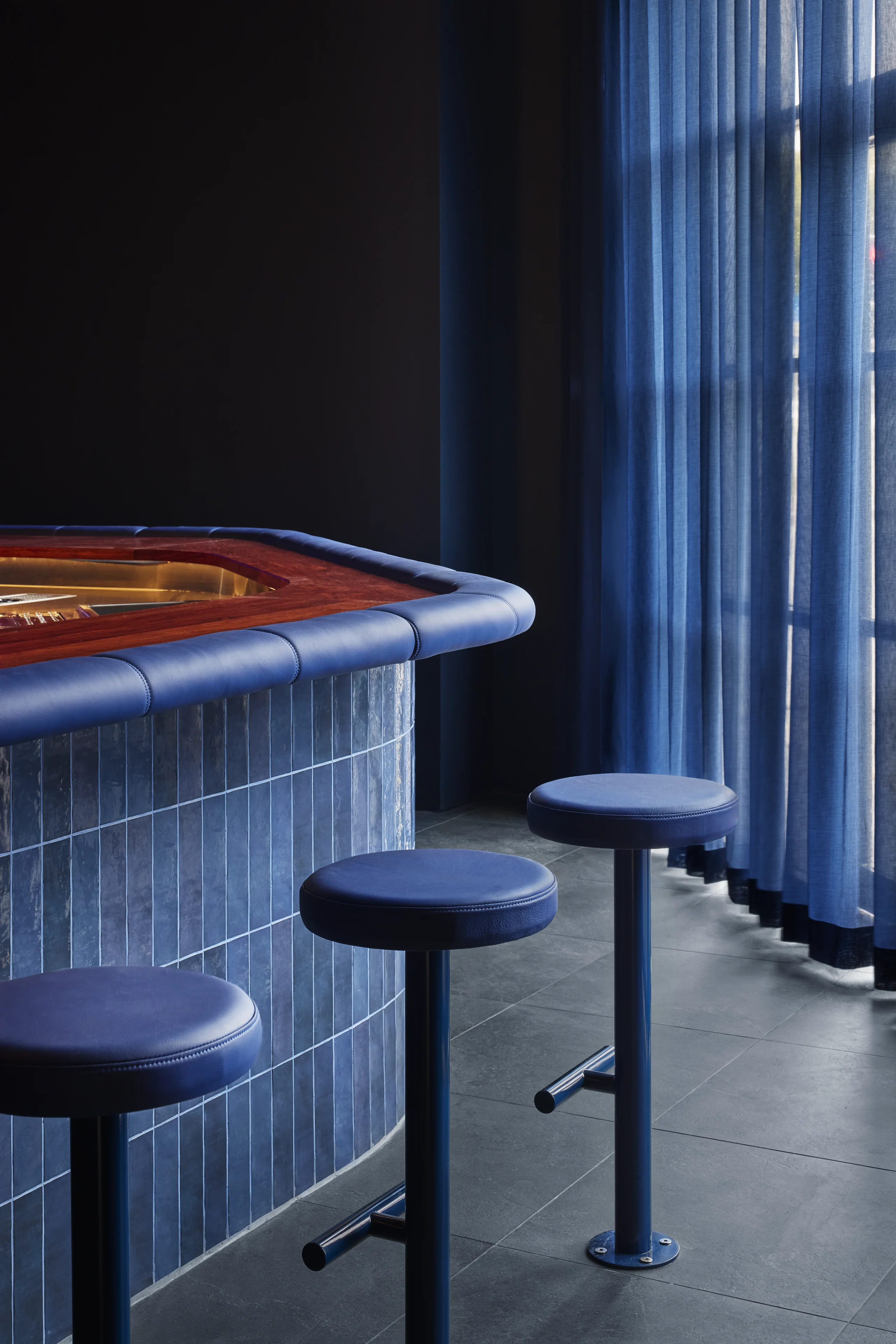 Blue cushioned bar stools lined along a blue tiled bar counter with blue curtain in background.