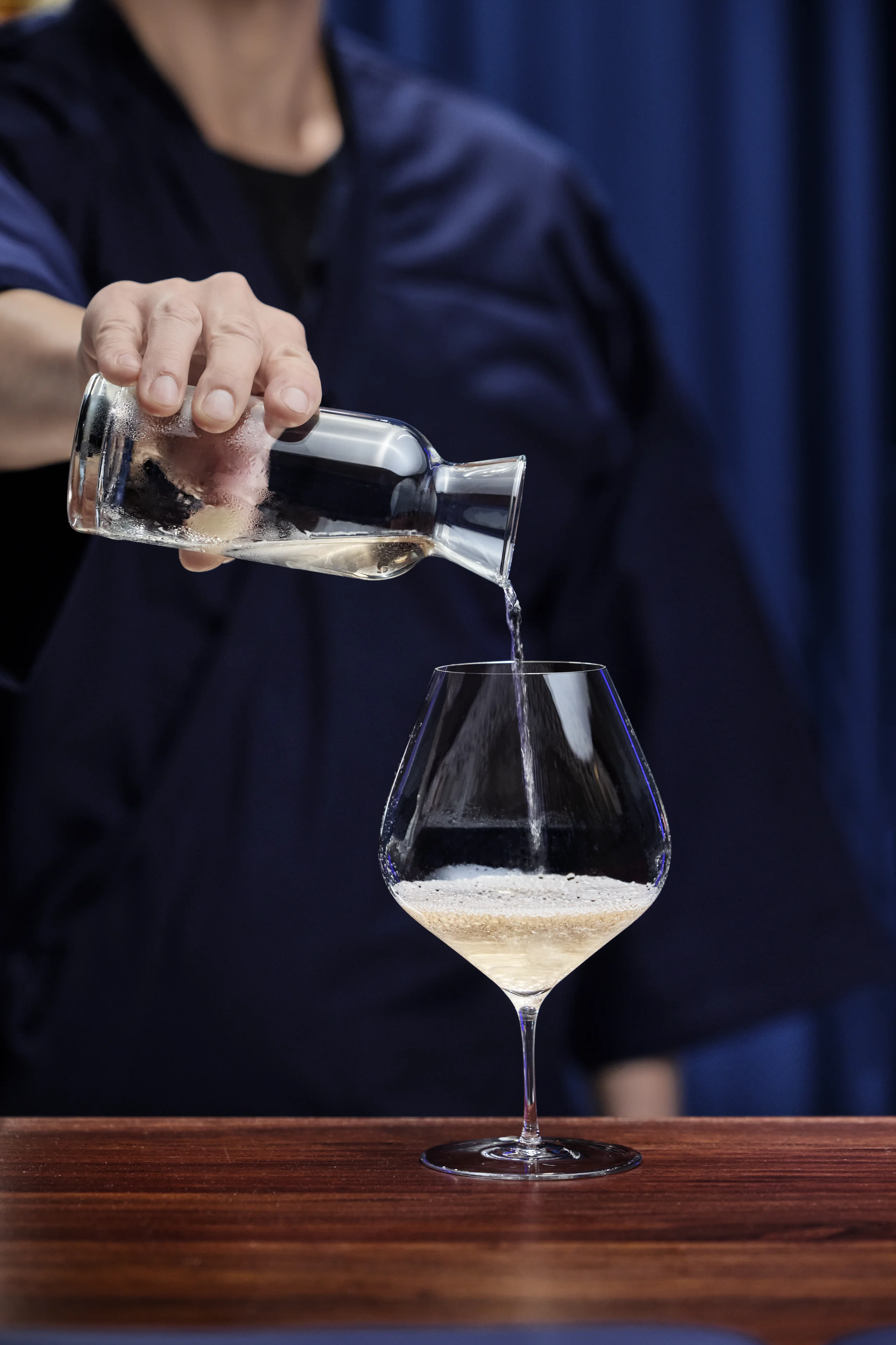 Person pouring a clear sparkling drink from a glass bottle into a wine glass on a wooden table.