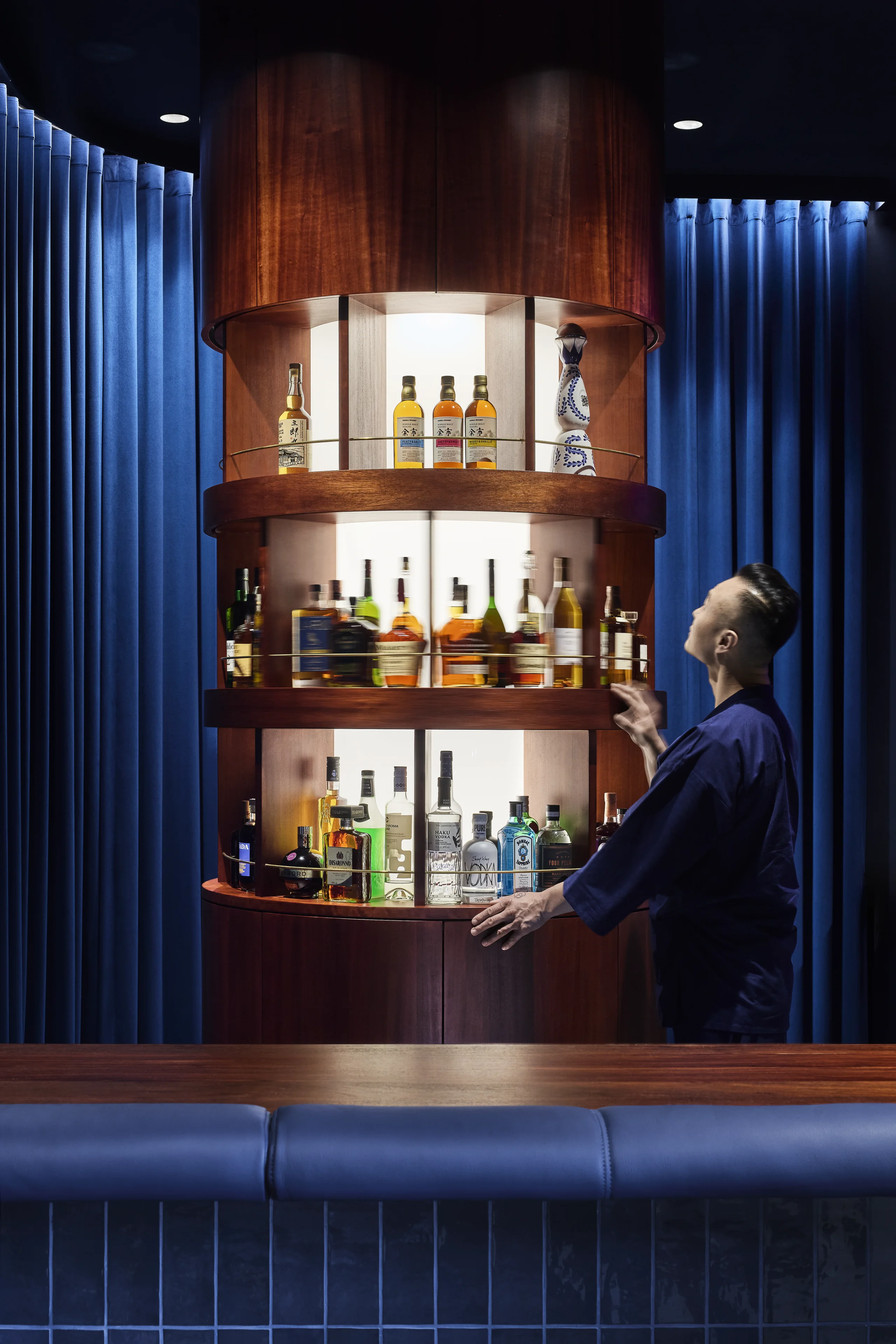 Man in dark clothing reaching for a bottle on a round wooden bar shelf filled with liquor bottles.