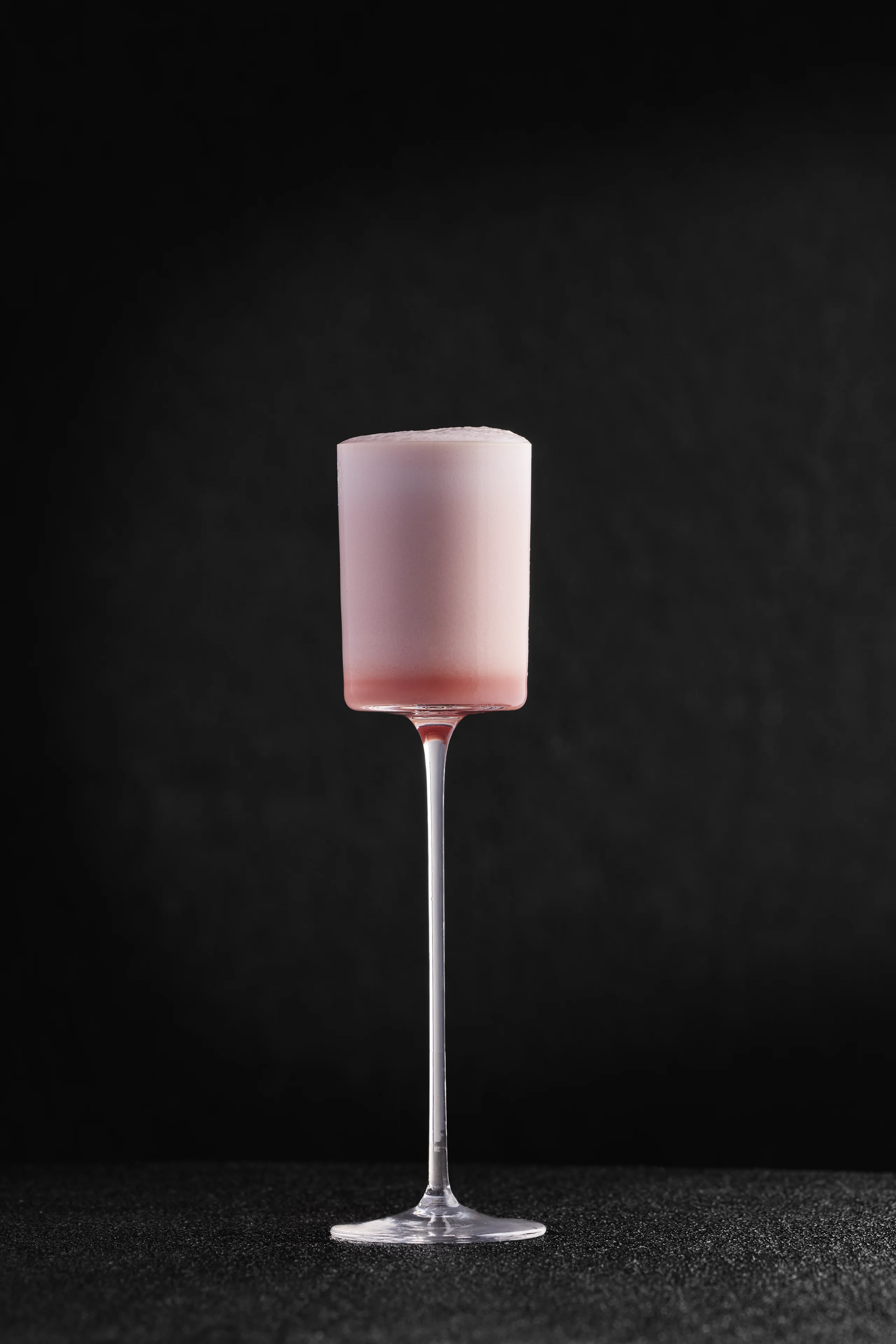 Tall glass with pink foam drink on a dark textured surface and black background.