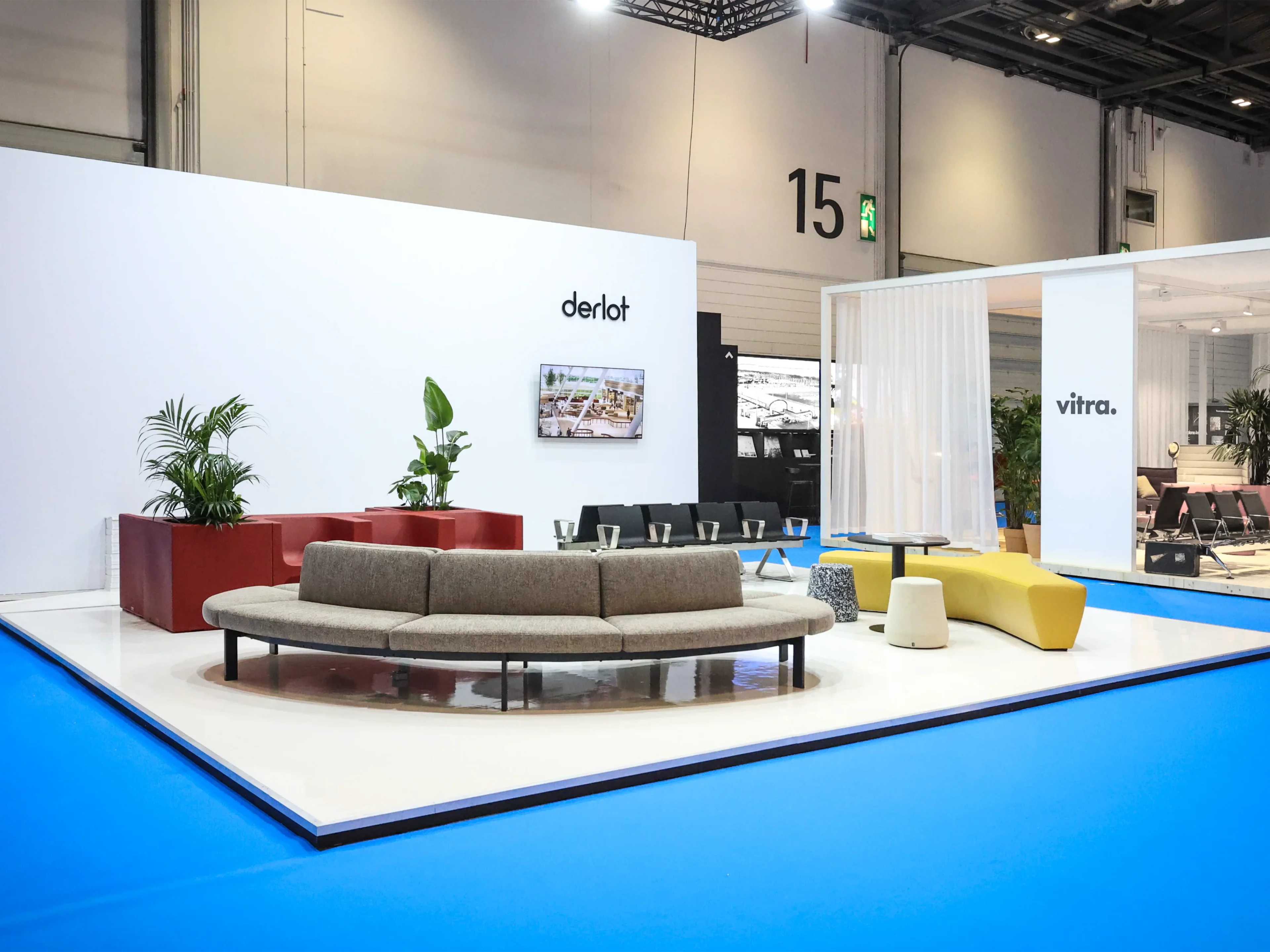 Modern furniture display including curved sofa, yellow bench, stools, and plants on white platform at trade show.