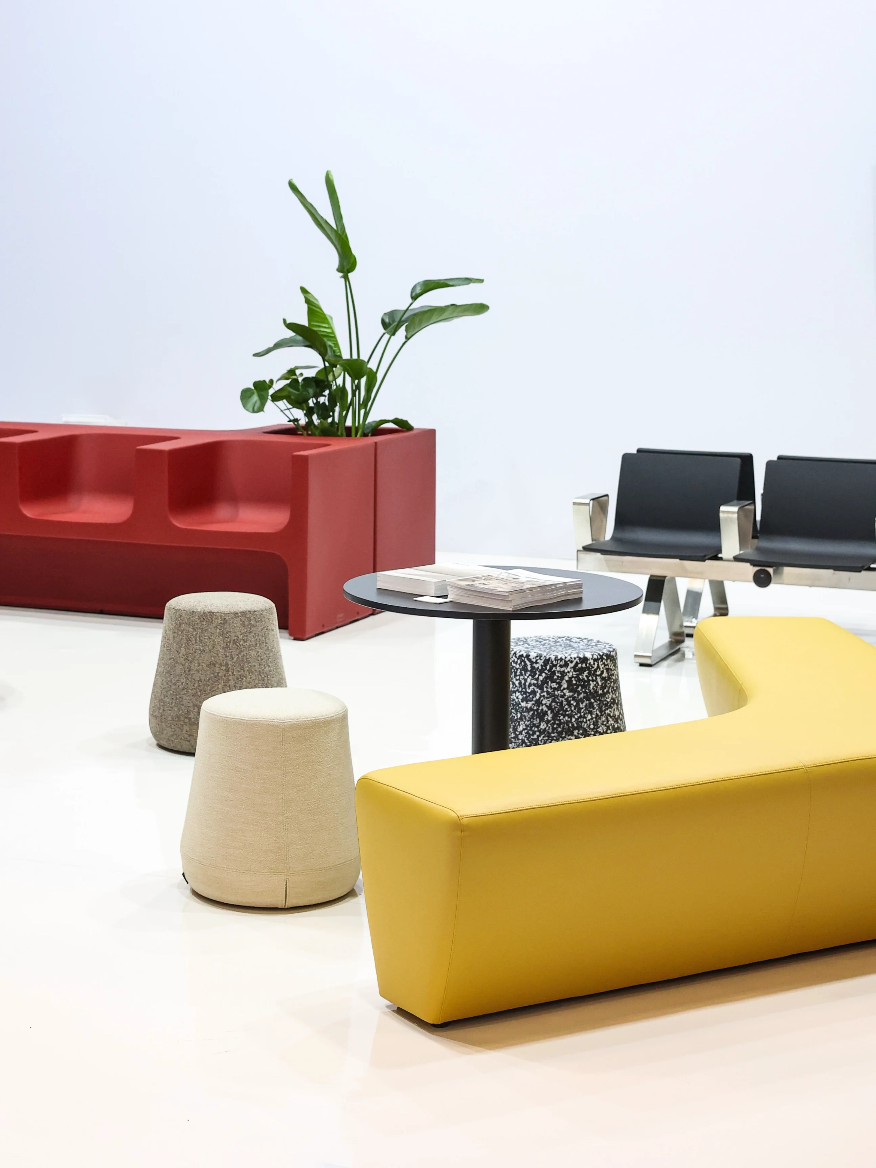 Modern waiting area with yellow sofa, round black table, multicolored stools, and red planter with green plant.