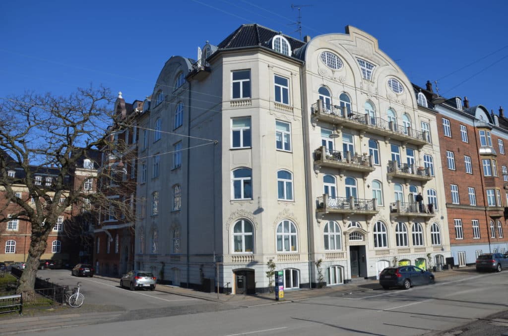 Hjørne på flere traditionelle europæiske lejlighedshuse på en byvej under en klar blå himmel.