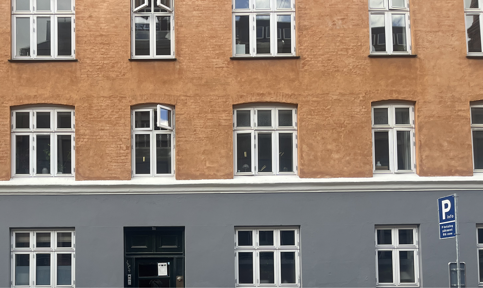 Ejendom med orangerøde murstensvægge og grå nederste facade med flere hvide vinduer og et enkelt åbent vindue. Repræsenterer andelsboliger som et vigtigt segment for Elby.