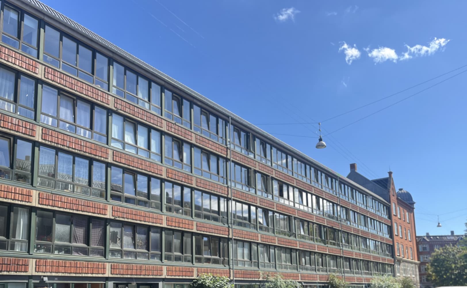 Gåvej langs en AAB-afdeling på Saxogade, Vesterbro,med sorte hegn, buske og parkerede biler under en klar blå himmel.