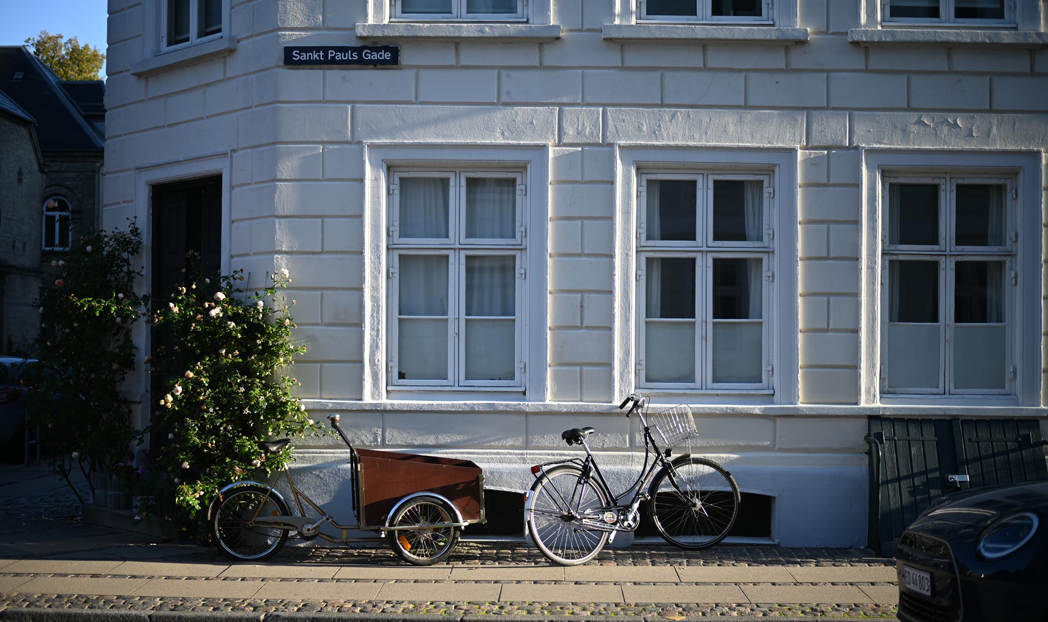 Sort ladcykel står parkeret foran et hvidt byhus med tre vinduer på en solrig dag på Sankt Pauls Gade. En typisk Elby gade