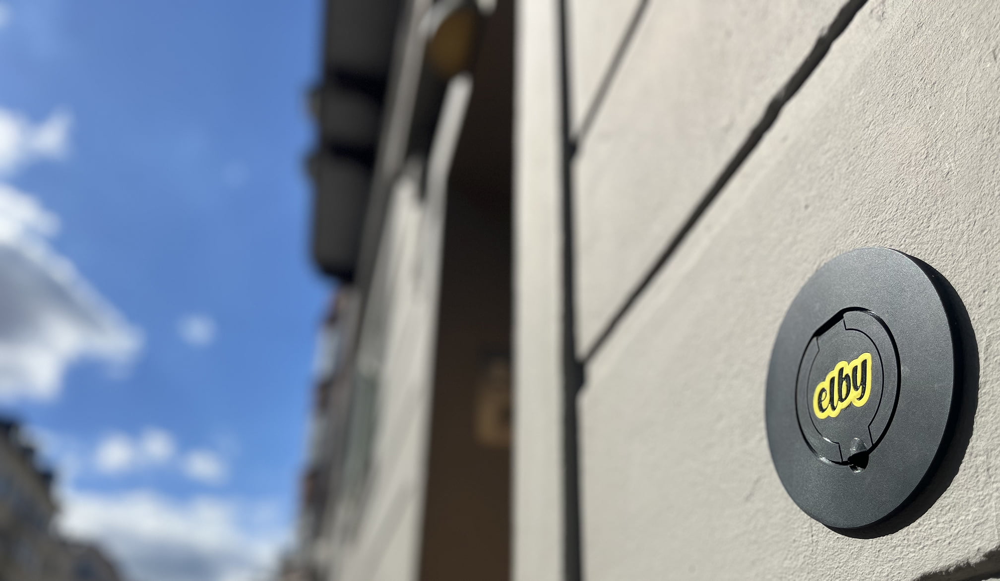 Sort væglader med gult 'elby'-logo monteret på en lys væg med blå himmel i baggrunden.