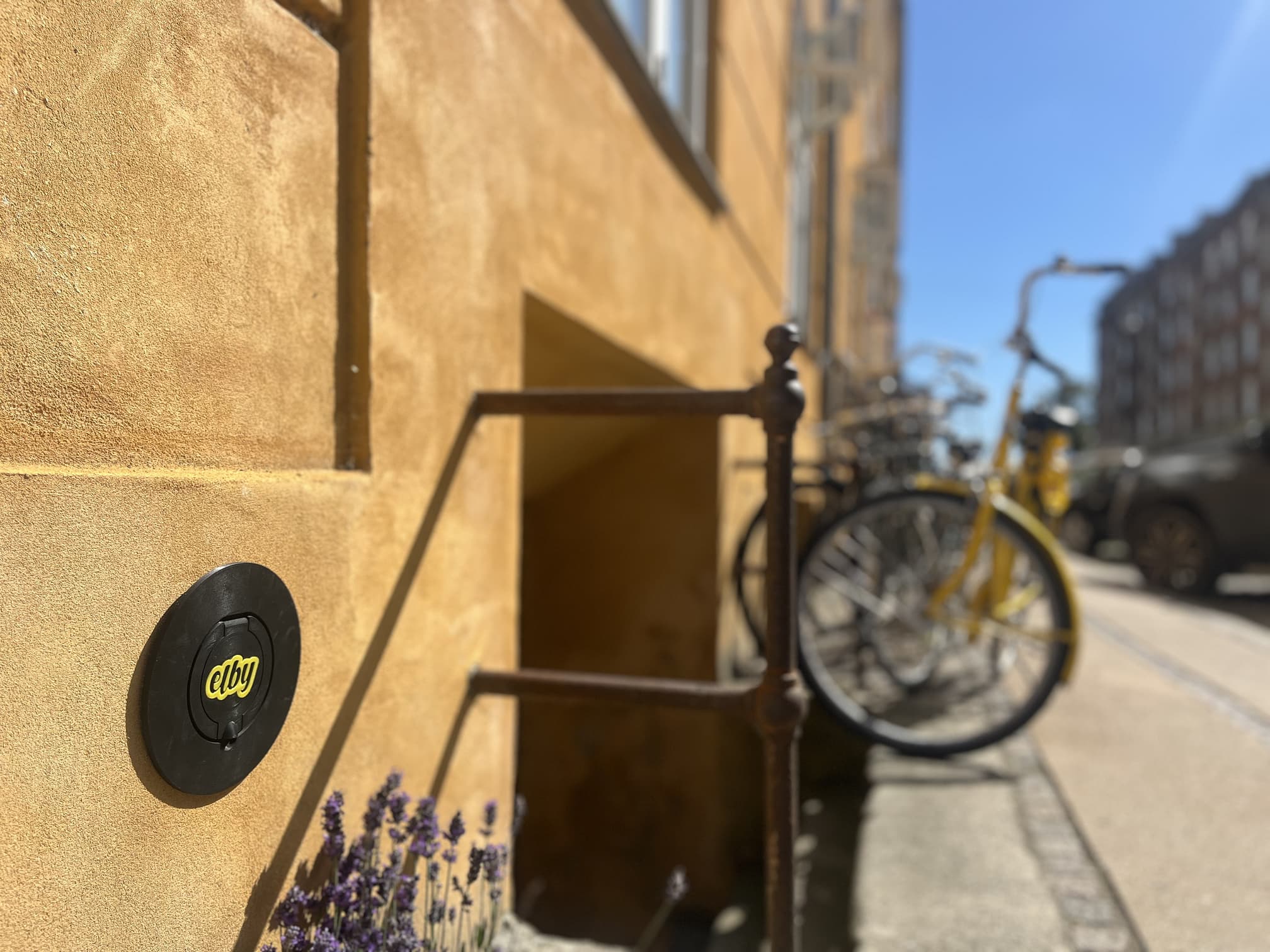 Sort cirkulært elbilslader mærket 'elby' monteret på en gul væg ved siden af en trappe med cykler i baggrunden på en solrig dag.