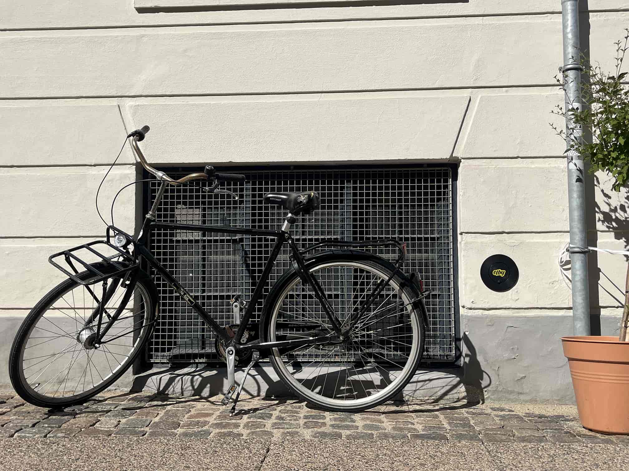Sort bycykel med kurv foran parkeret mod en hvid mur med gitternet vindue og en plante i en orange potte på fortovet. Elby væglader monteret i facaden.
