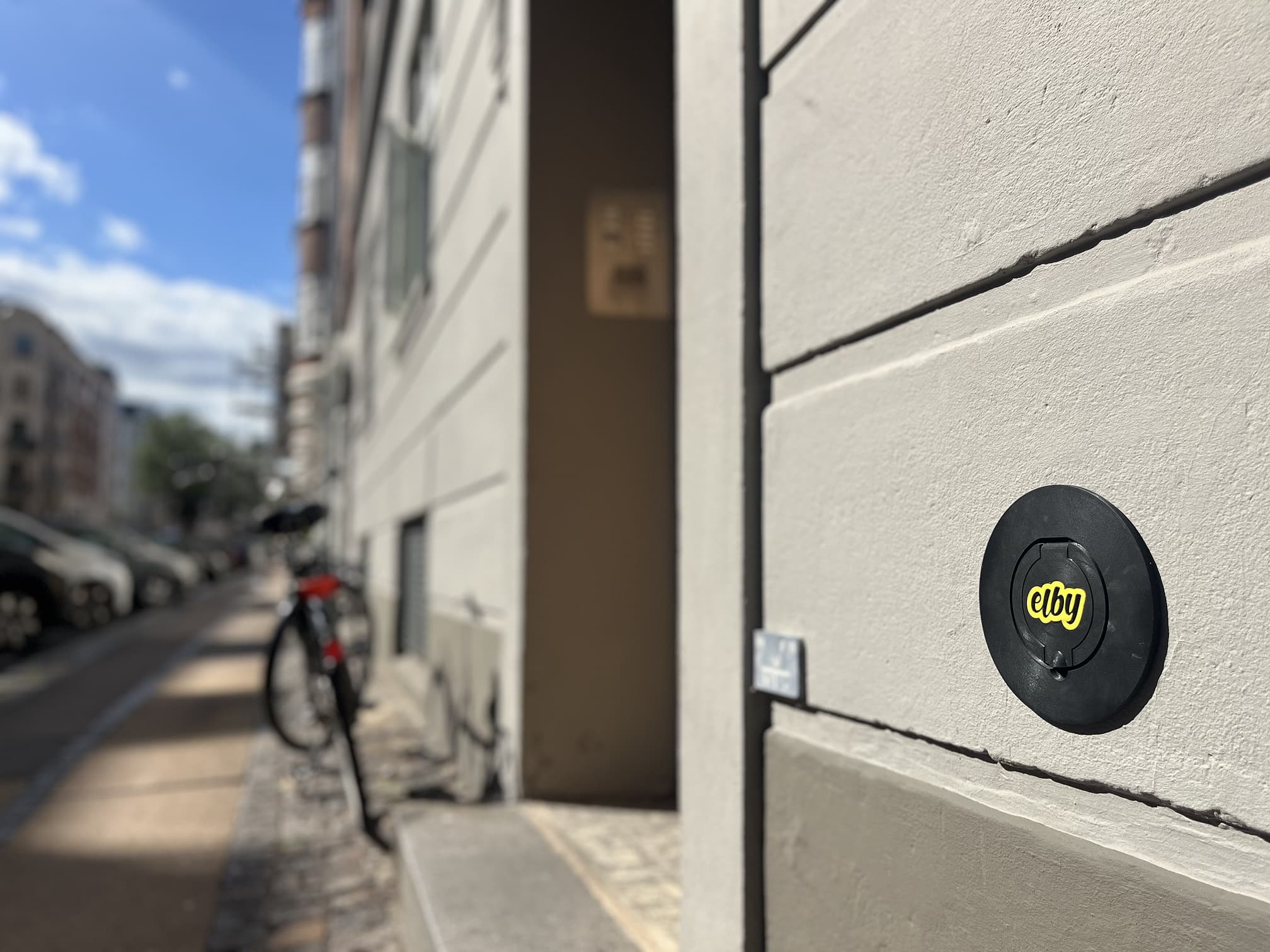 Sort cirkulært stik med logoet 'elby' på en hvid væg i en bygade under solskin.