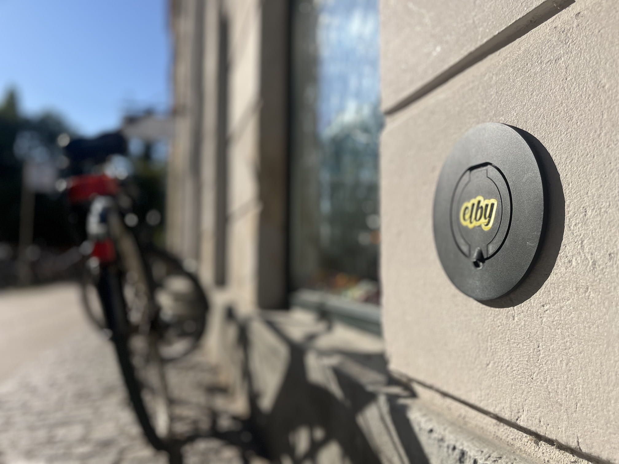 Sort rund skilt med teksten 'elby' monteret på en byggevæg med en sløret cykel i baggrunden.
