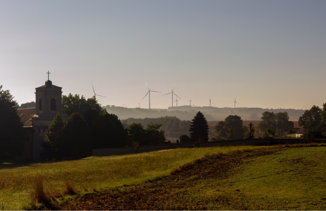 Kostel s křížem na věži vedle stromů a pole s větrnými turbínami na obzoru za mlhavého dne.