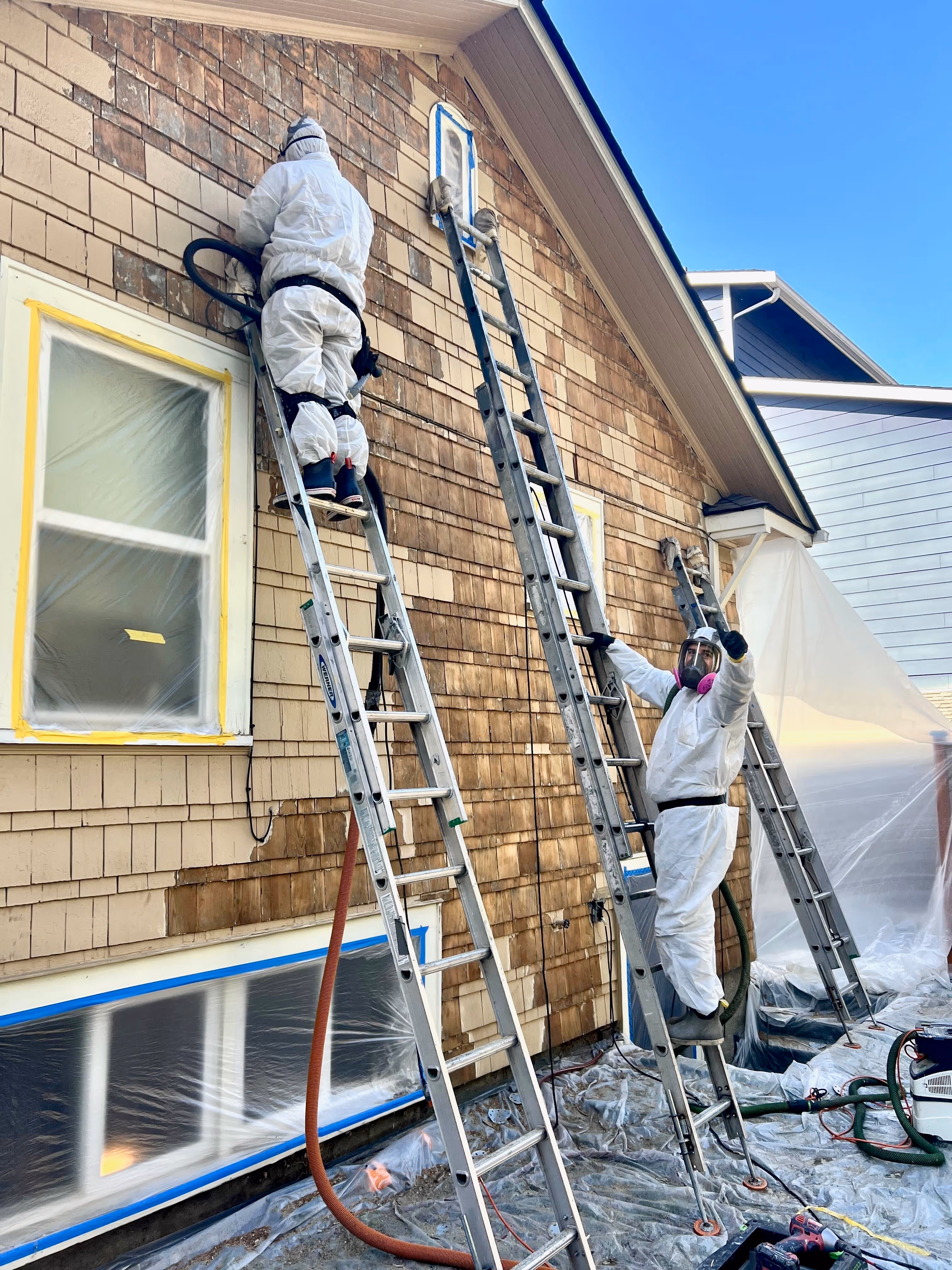Two workers in protective suits on ladders stripping wood siding from a house exterior.