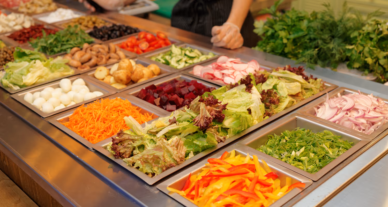 Salad bar with fresh ingredients including lettuce, shredded carrots, sliced radishes, cucumbers, cherry tomatoes, red onions, and assorted greens in metal containers.