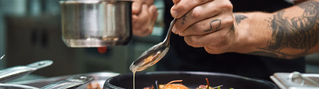 Chef with tattooed arm drizzling sauce from a spoon onto a plated dish in a kitchen.