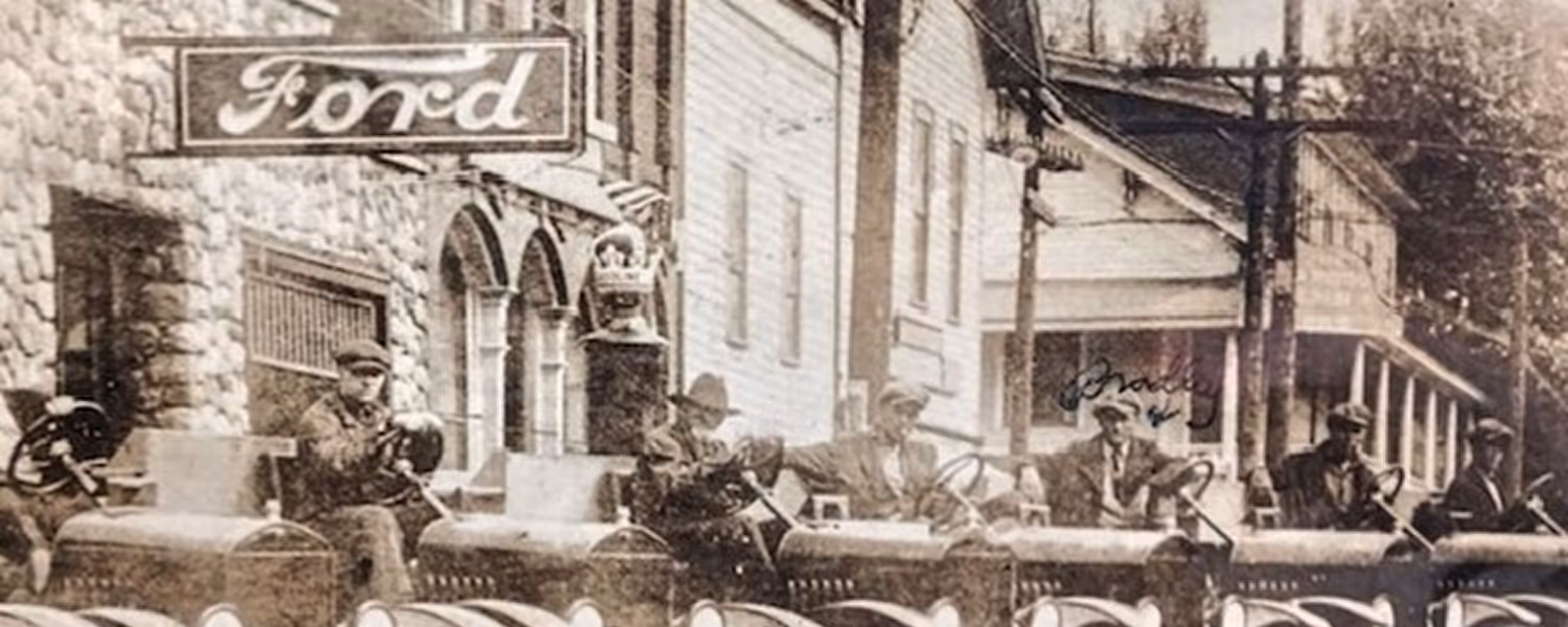 Sepia-toned historical photo showing several men sitting in early Ford automobiles parked in a row outside a stone building with a Ford sign in Clarktson