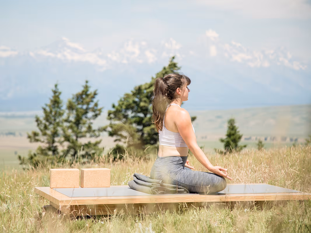 Woman in athletic wear meditating in a cross-legged yoga pose outdoors on a wooden platform with greenery and mountains in the background.